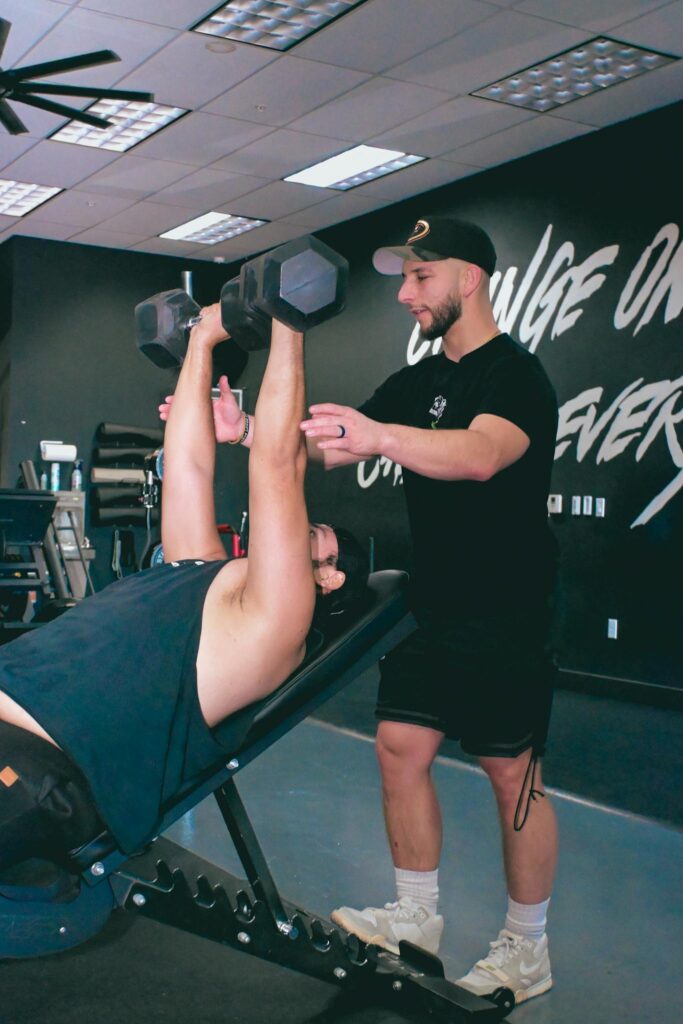 Trainer helps a person during a dumbbell exercise at a gym, emphasizing proper form and safety.